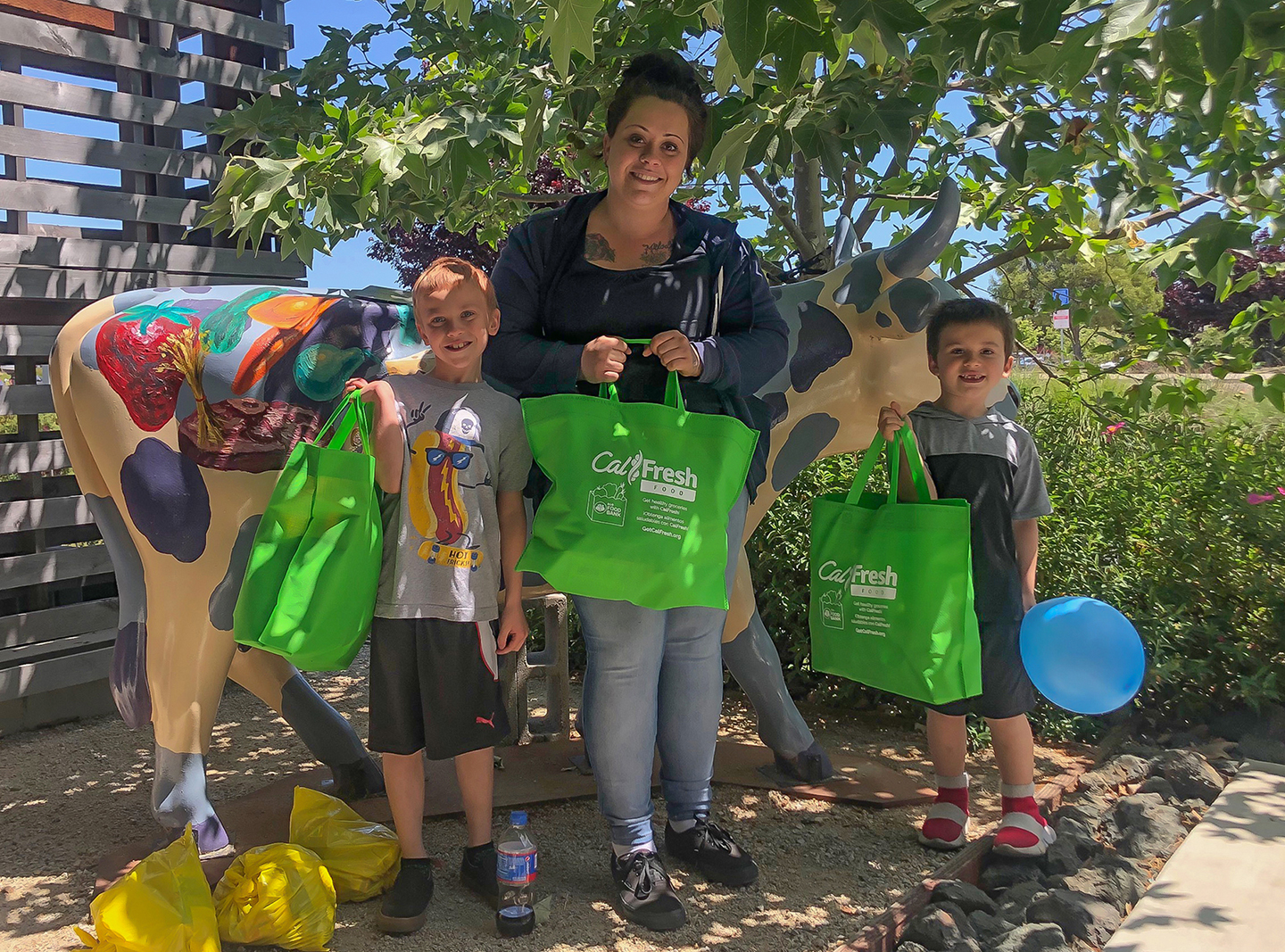 A woman and two children hold green CalFresh Food Benefits bags next to a painted cow sculpture outdoors.